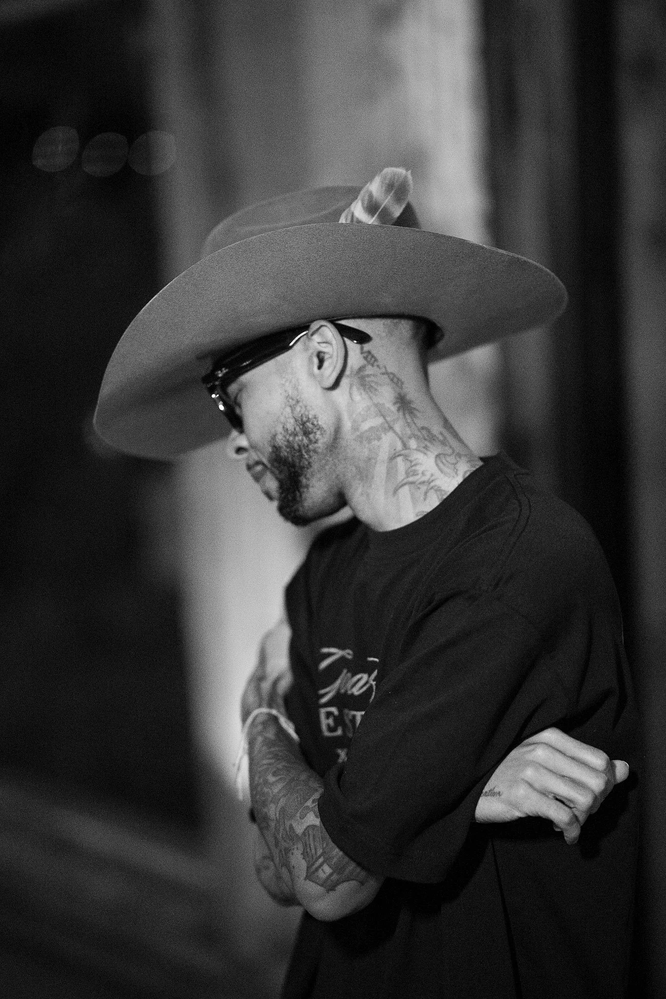 A man with tattoos on his neck and arms, wearing sunglasses and a cowboy hat, standing with crossed arms.