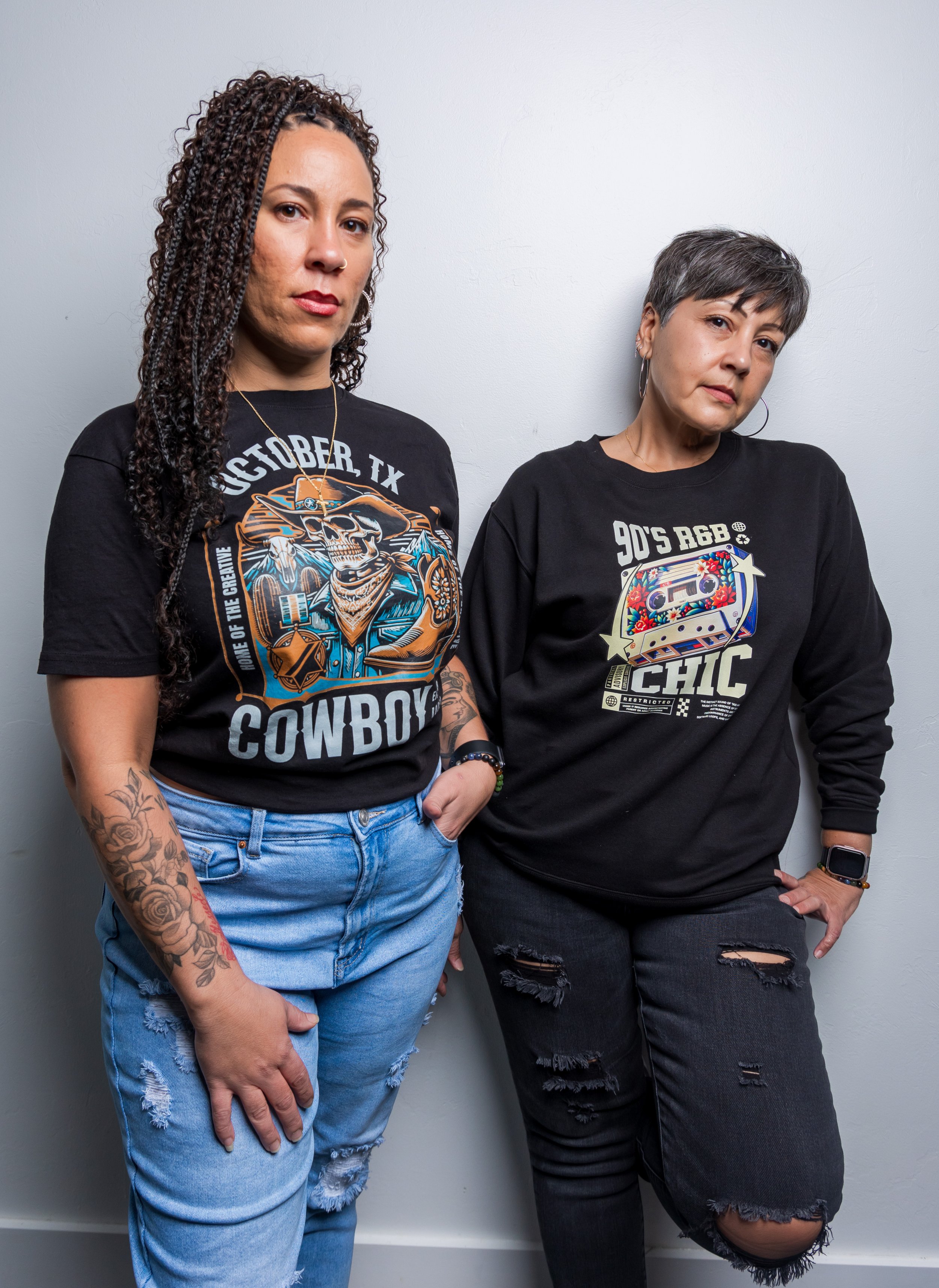Two women stand against a plain gray wall. The woman on the left has curly hair, tattoos on her arm, and wears a black T-shirt with a skeleton graphic and the words 'OCTOBER TX COWBOY.' The woman on the right has short, gray hair, wears a black T-shirt with a graphic featuring a cassette tape and the words '90'S R&B,' and black ripped jeans. They both have neutral expressions and pose casually.