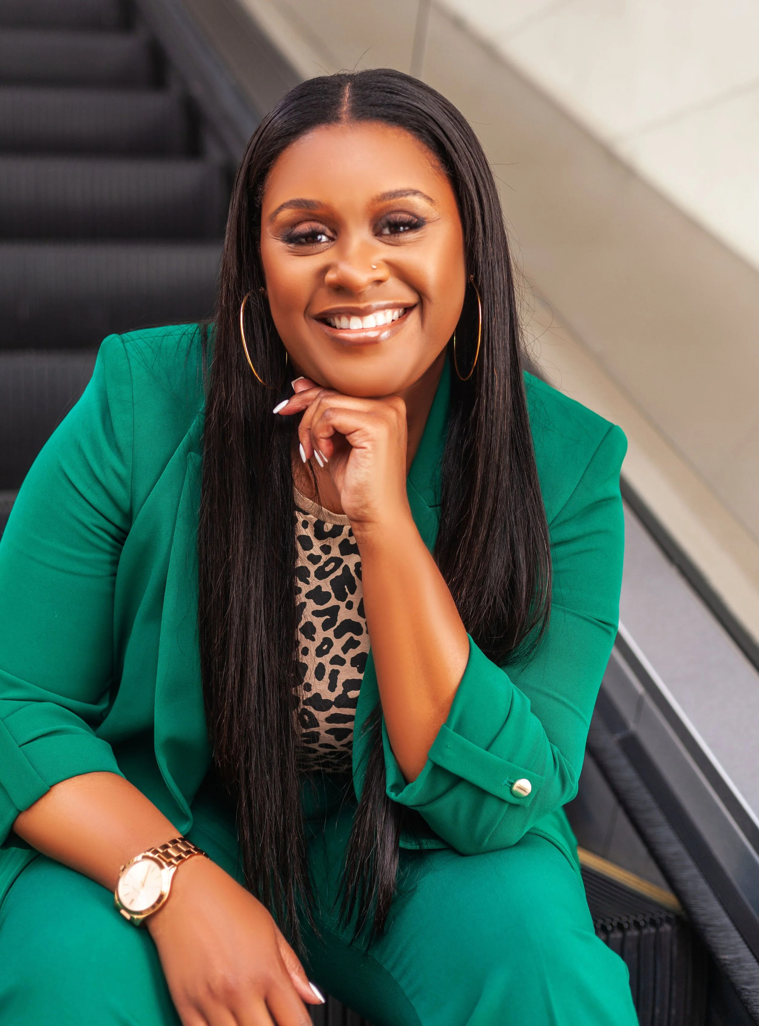 A woman with dark hair styled straight, wearing a green blazer and matching green pants, gold hoop earrings, a gold watch, and a leopard print top underneath, sitting on an escalator with a smile, resting her chin on her hand.