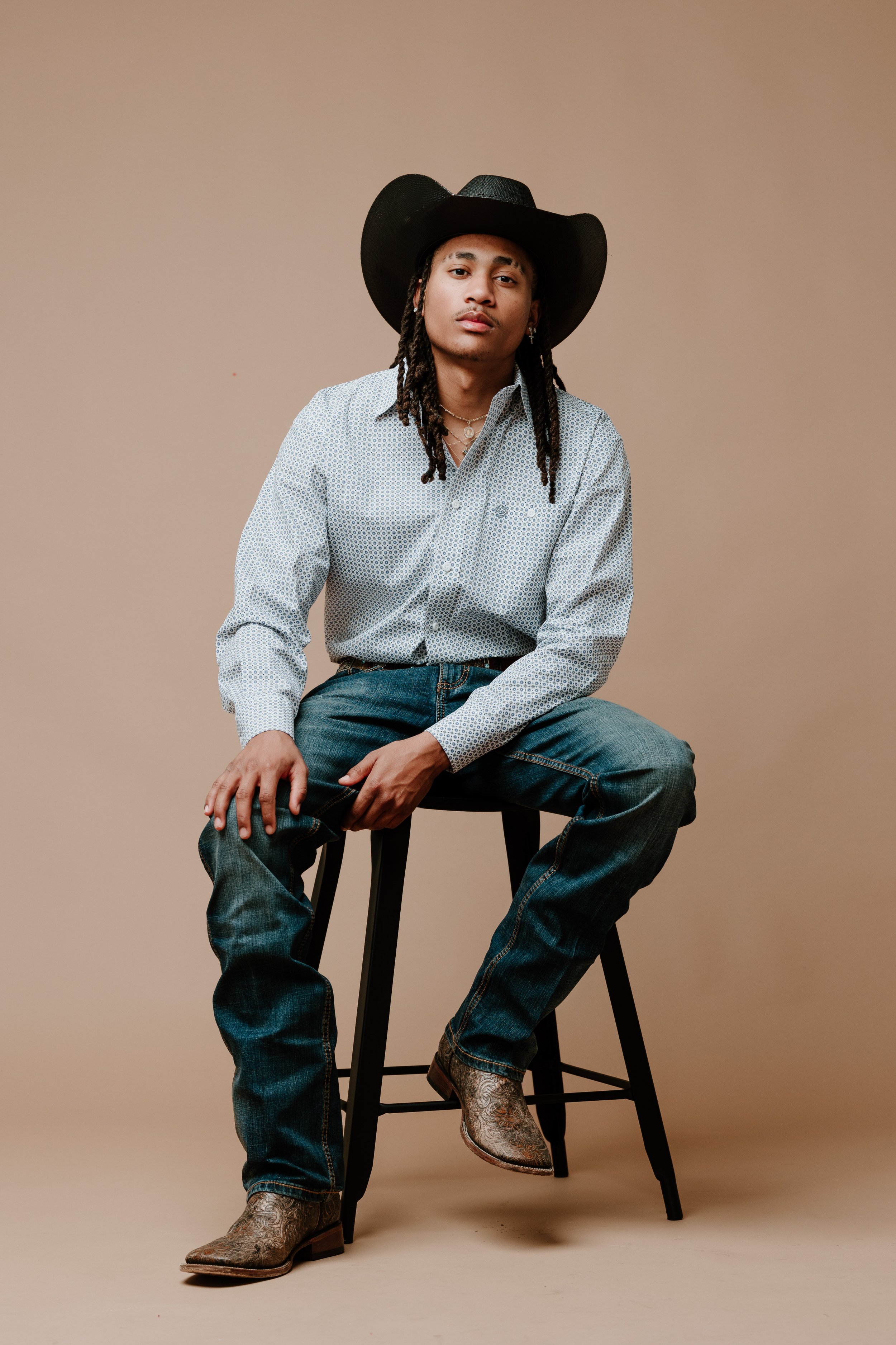 A young man with long braided hair wearing a cowboy hat, a patterned button-up shirt, jeans, and cowboy boots sitting on a stool against a beige background.
