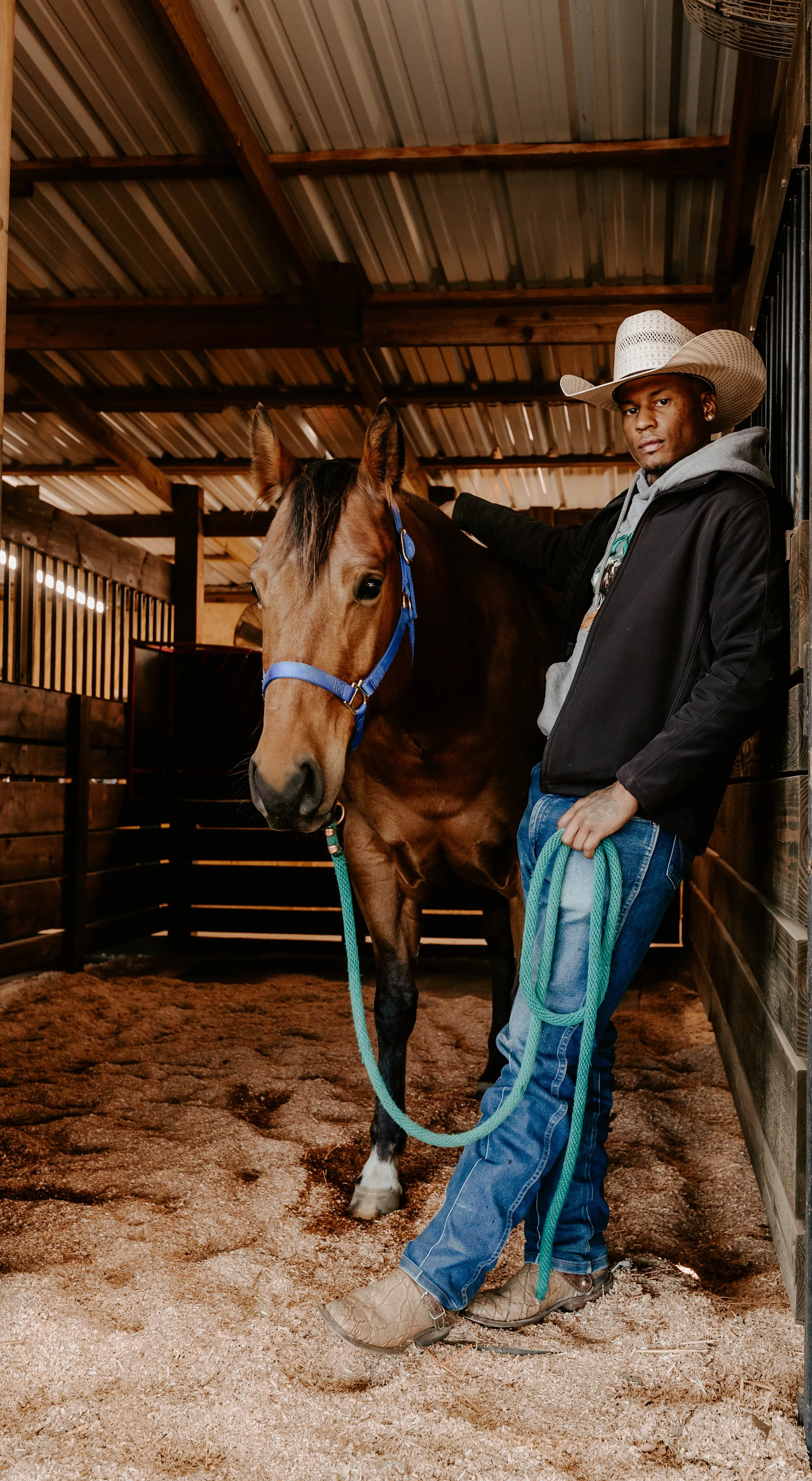 A man wearing a cowboy hat, black jacket, and jeans stands next to a brown horse with a blue halter inside a wooden stable. The man has his hand on the horse's neck and is holding a green lead rope.