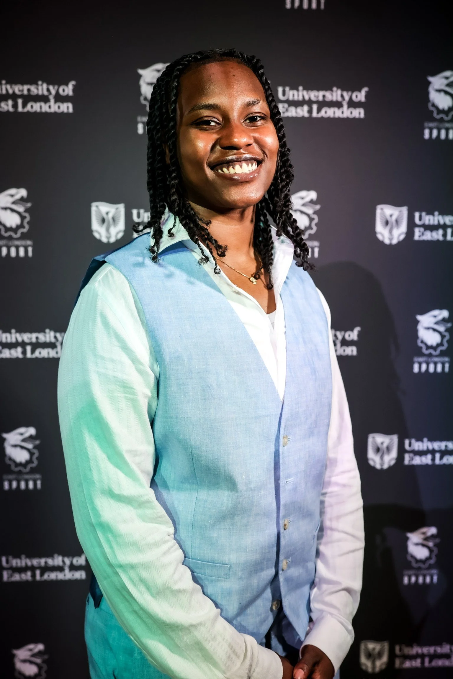 Smiling woman with twisted hair wearing a light blue and white vest, standing in front of a black background with the logos and text of the University of East London and sports branding.