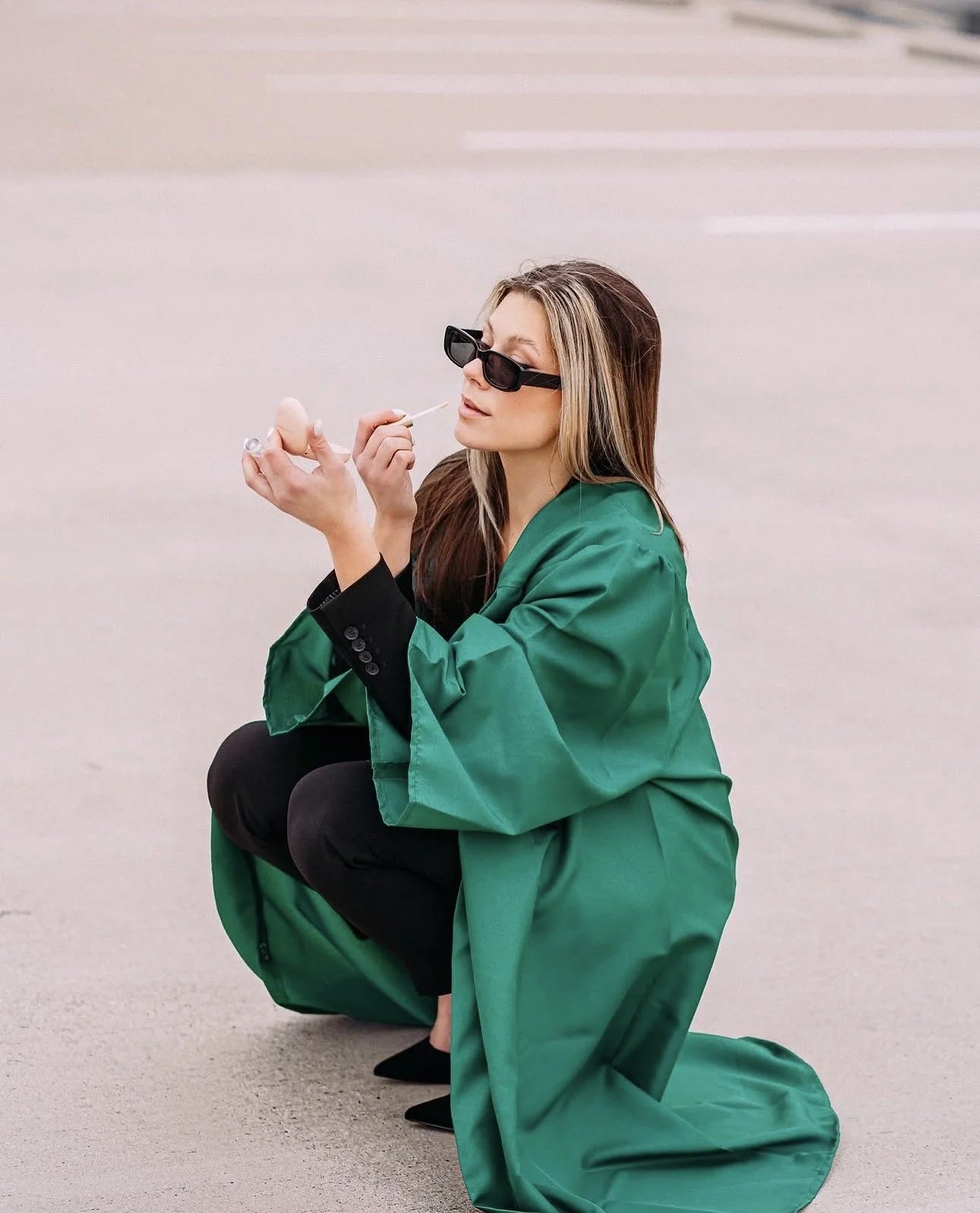 Woman in sunglasses and a green suit crouching on a light-colored surface, holding a cigarette and looking to the side.