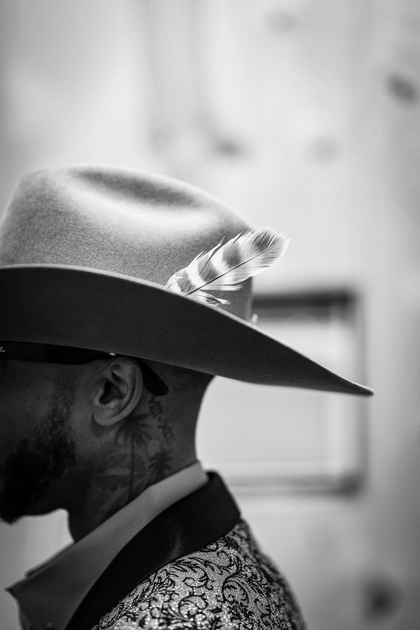 Side profile of a person wearing an elaborate hat decorated with a feather, with lace gloves, and visible tattoos on the neck and behind the ear, in black and white.