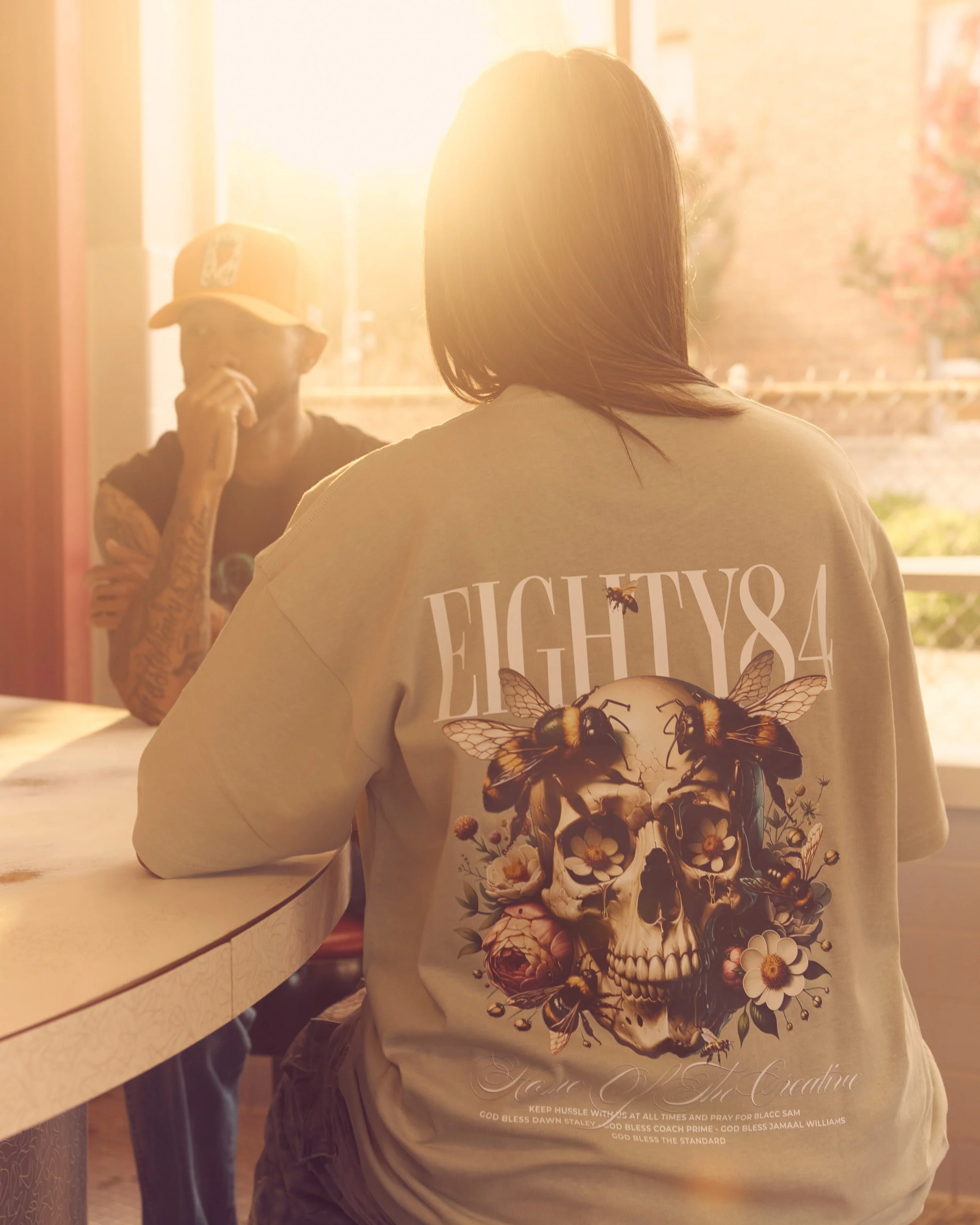 A woman and a man inside a room with sunlight streaming through the window. The woman has shoulder-length hair and is wearing a beige T-shirt with a skull and floral design on the back. The man is wearing a baseball cap and sunglasses, with tattoos on his arms, and is talking on the phone.