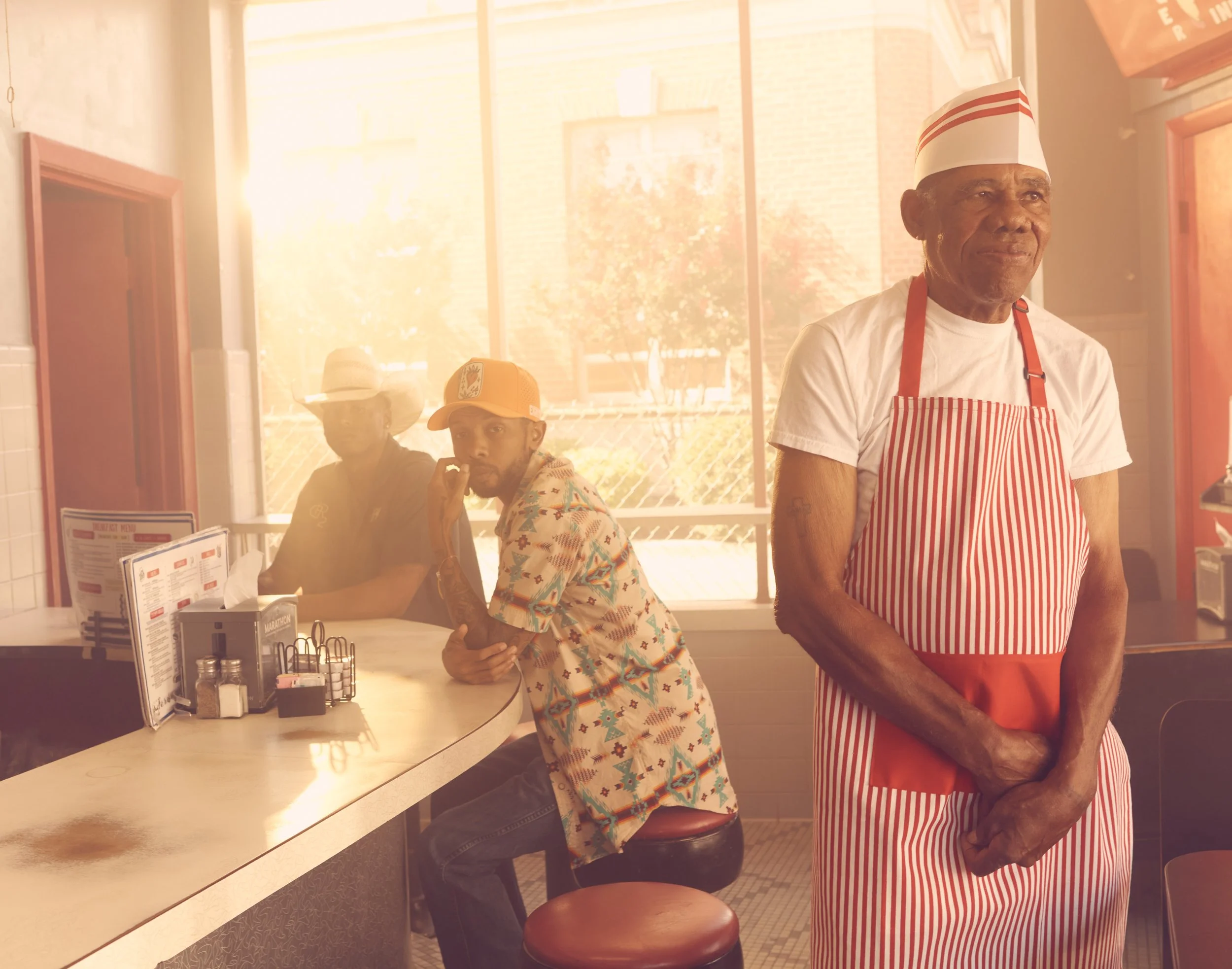 A cafe or diner scene with three men: one older man in a red and white striped apron and hat standing near the counter, and two younger men sitting at the counter, one wearing a colorful patterned shirt and a hat, the other wearing a hat and a patterned shirt, all inside with large windows letting in natural light.