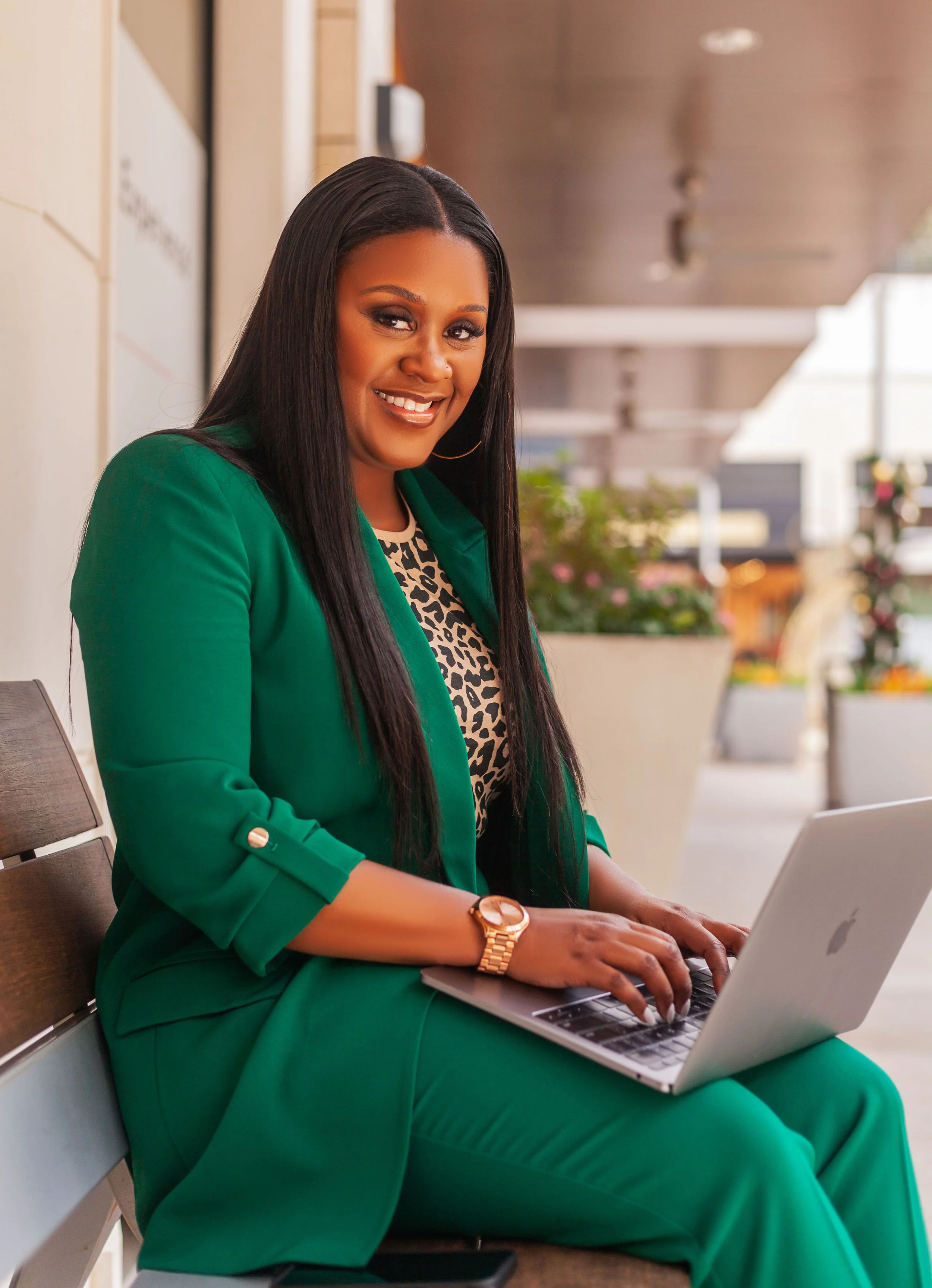 A woman with long black hair, wearing a green blazer and pants, and a leopard print top, sitting on a wooden bench at an outdoor location, working on a silver laptop, smiling.