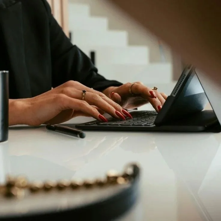 Close-up of a woman’s hands typing on a compact keyboard, with her fingernails painted red, sitting at a white desk.