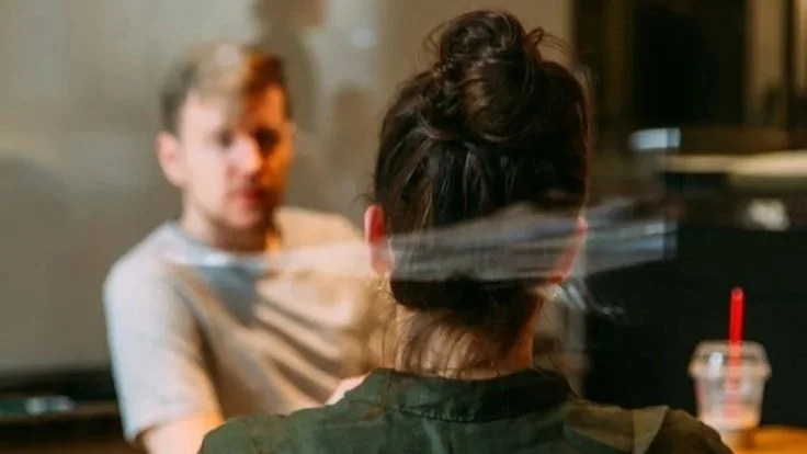 A woman with her hair in a bun facing a young man in a casual setting, with a drink in a plastic cup with a red straw on the table.