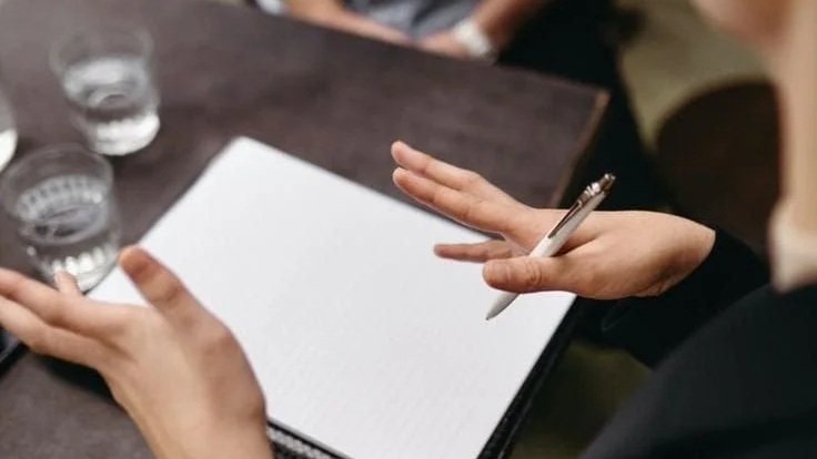 A person holding a pen above an empty notebook on a table with three glasses of water.