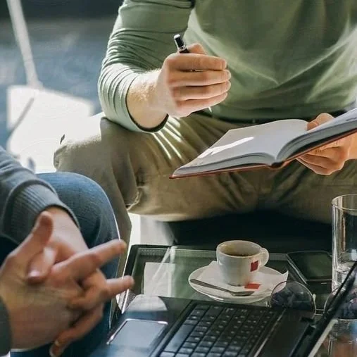 Person taking notes in a notebook during a meeting, with a coffee cup, laptop, and smartphone on the table.
