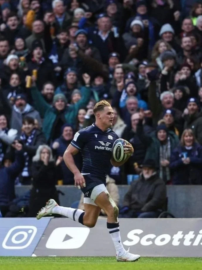 A rugby player running with a ball in hand during a match, with a large crowd of spectators cheering in the background.