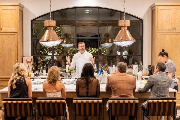 A chef giving a cooking demonstration or class in a kitchen with a group of six people seated at a counter, with large window and pendant lights above.