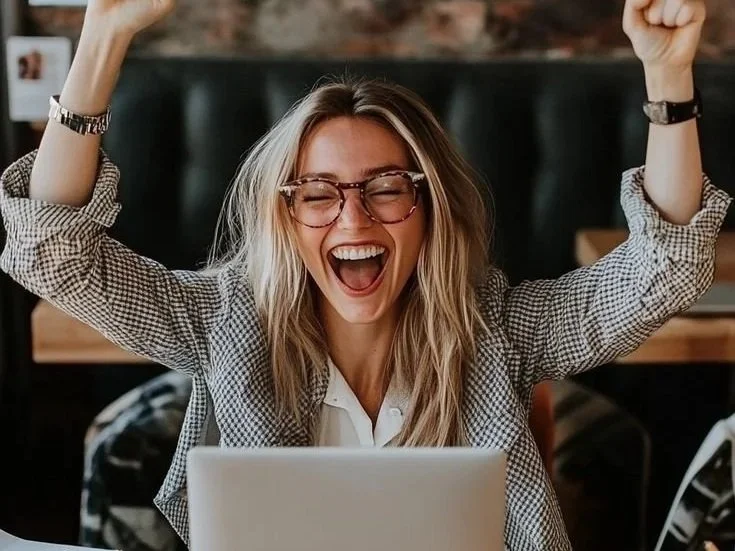 A woman with glasses and blonde hair celebrating joyfully in front of a laptop.