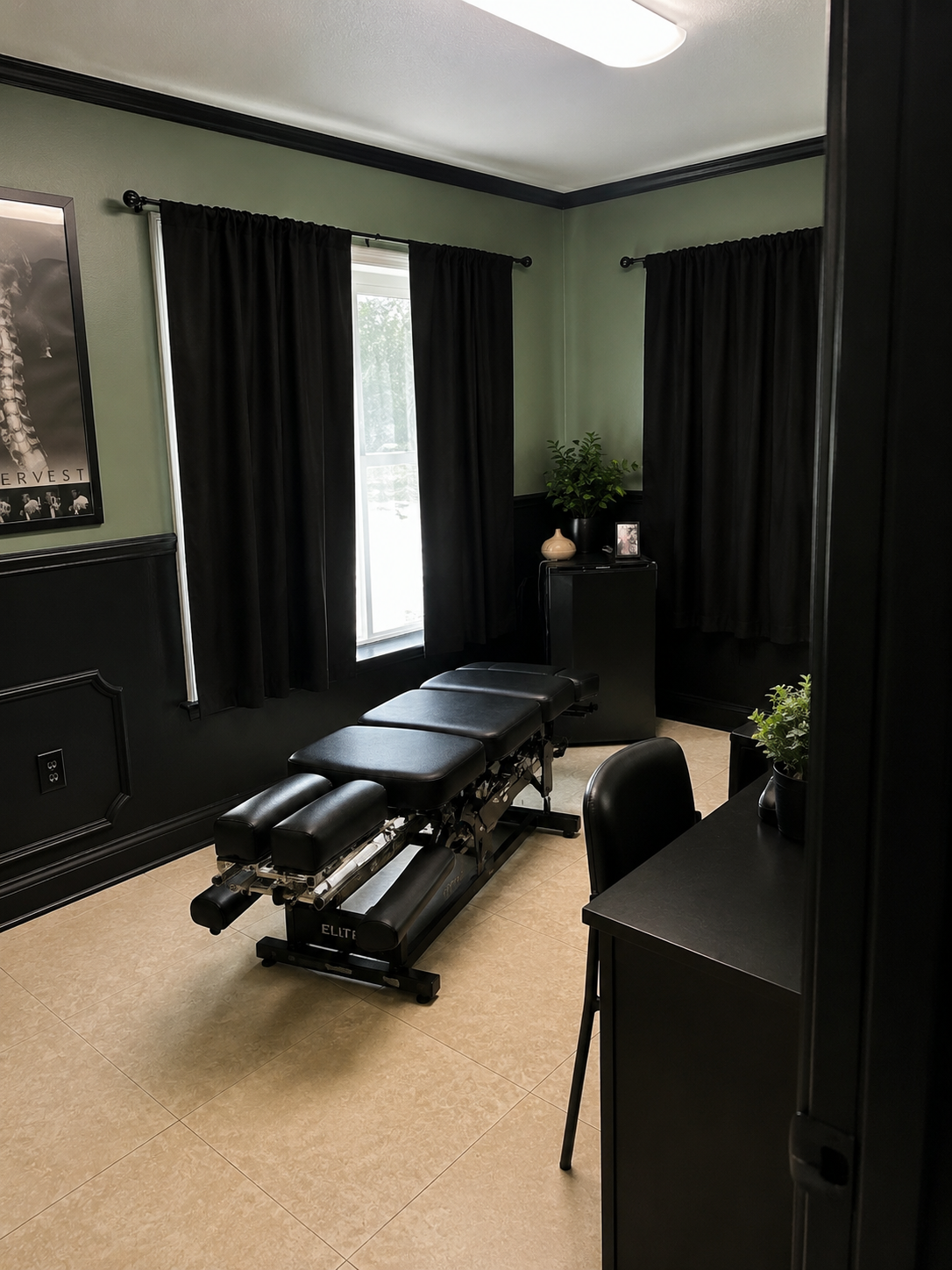 A massage therapy room with a black massage table, green walls, black curtains, and plants on a small cabinet and a desk.