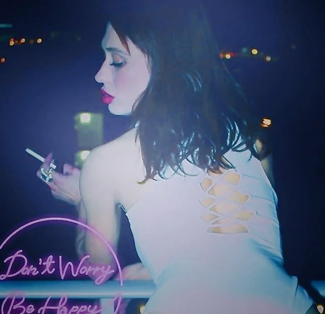 A woman with dark hair and makeup, wearing a white dress with lace-up back, looks down at her phone against a city nightscape background.
