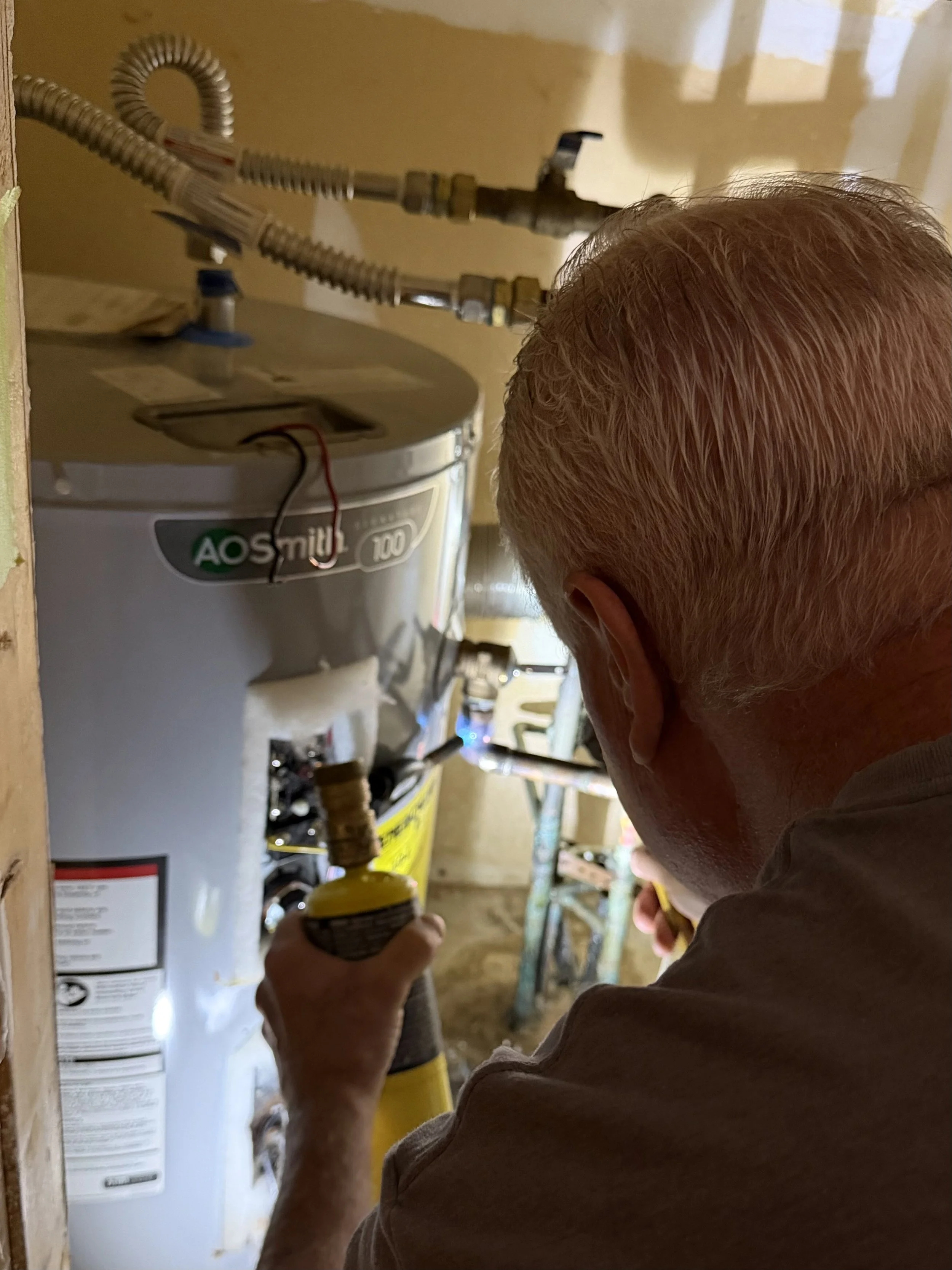 A person with short, gray hair working on a plumbing task in a garage or workshop, using a yellow screwdriver near a water heater or boiler, with tools and supplies in the background.