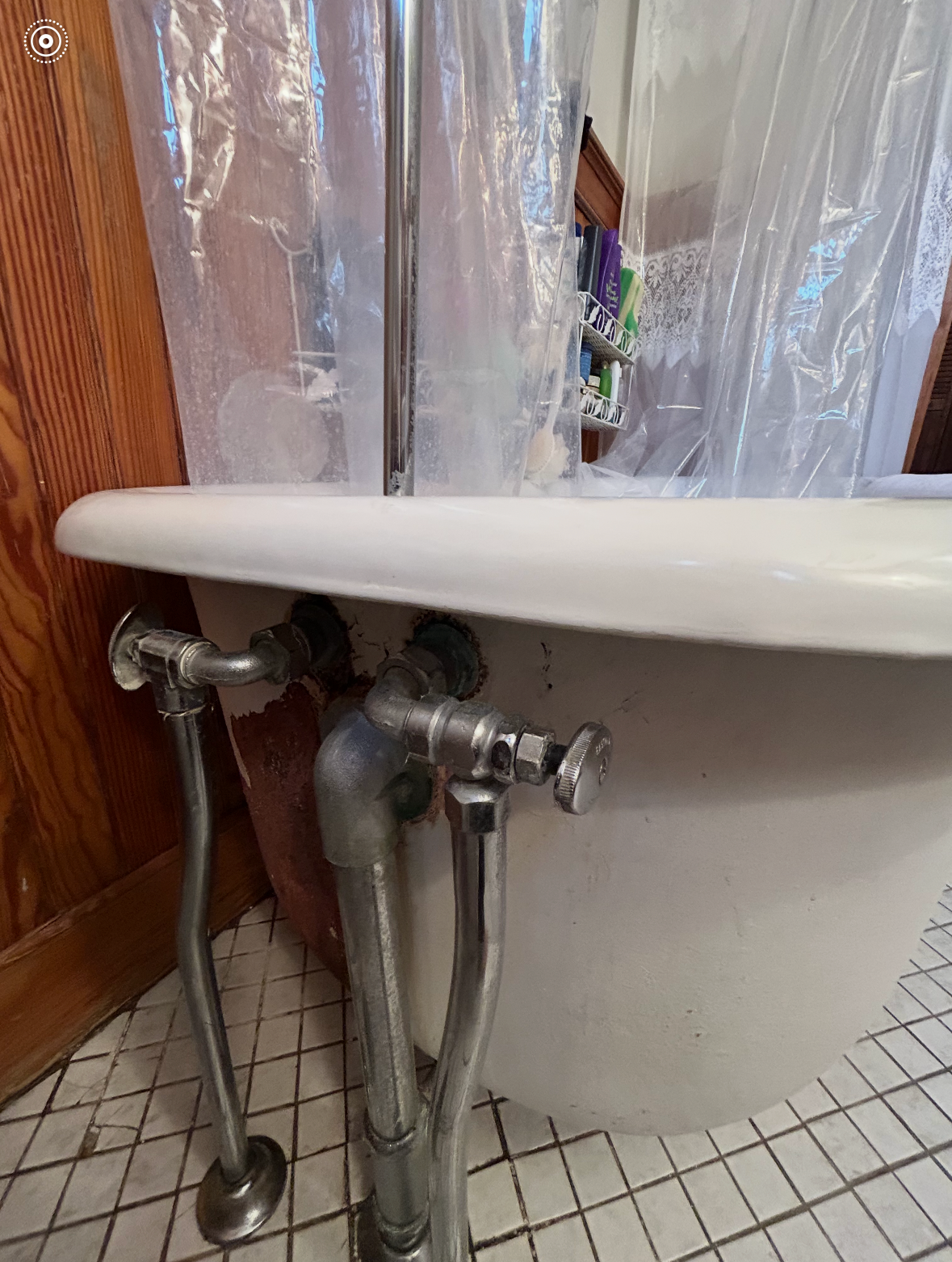 Close-up of an old bathtub with exposed plumbing, shower curtain, and tiled bathroom floor.