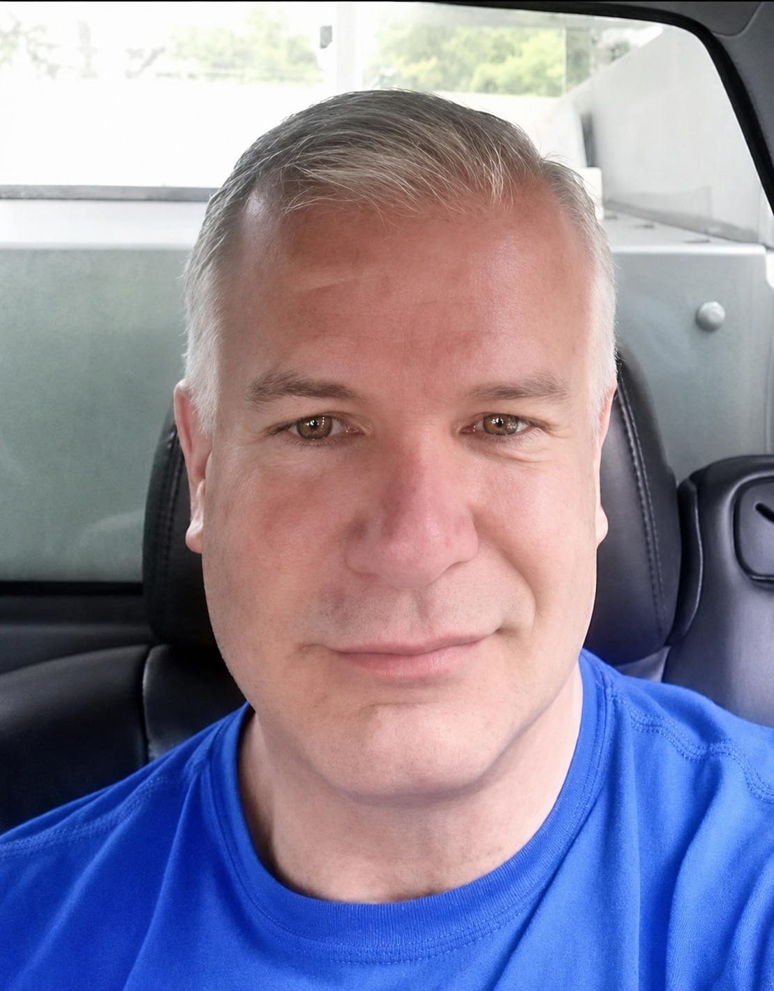 A man with short gray hair smiling while sitting in a vehicle with a black leather seat back and a window showing a cloudy sky.