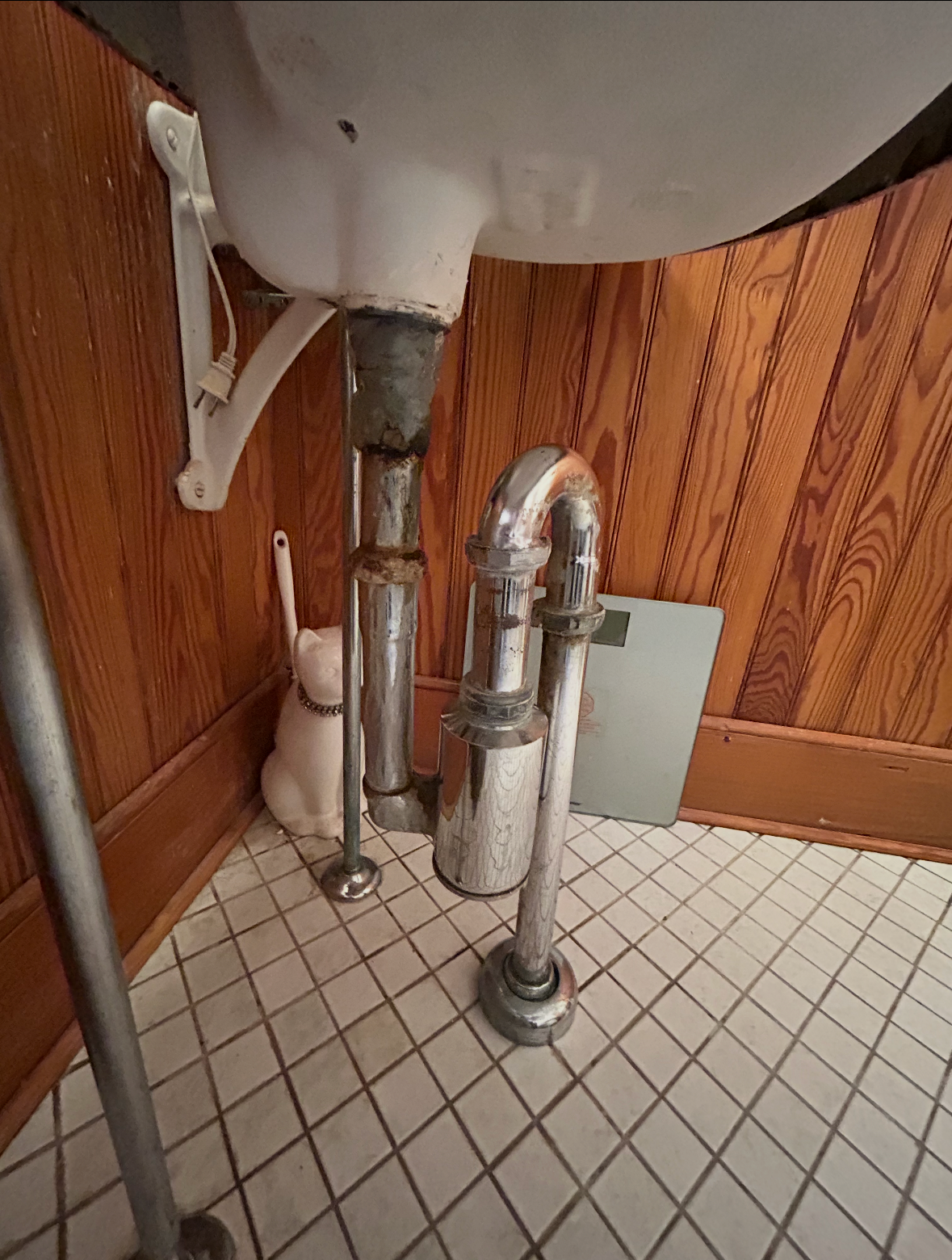 Under the sink pipes and plumbing with a wood-paneled wall and tiled floor in the background.