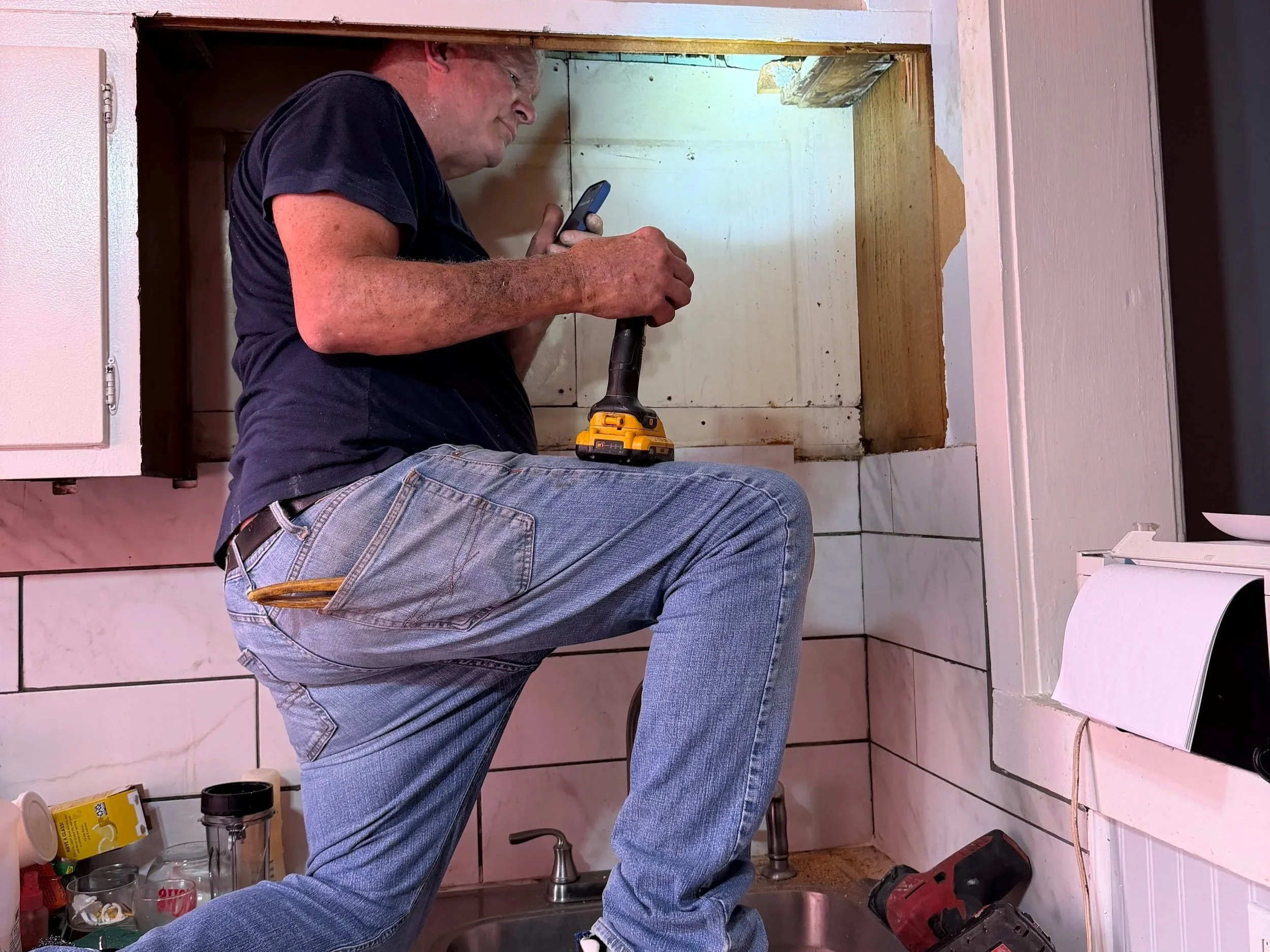 A man wearing a black t-shirt and blue jeans is kneeling on the floor and working on a small kitchen cabinet, holding a cordless drill. The space inside the cabinet is empty, with wood trim visible along the top edge. Various tools and containers are on the counter below him, including a cup and some bottles. The room has tiled walls and a white appliance or cabinet to the right.