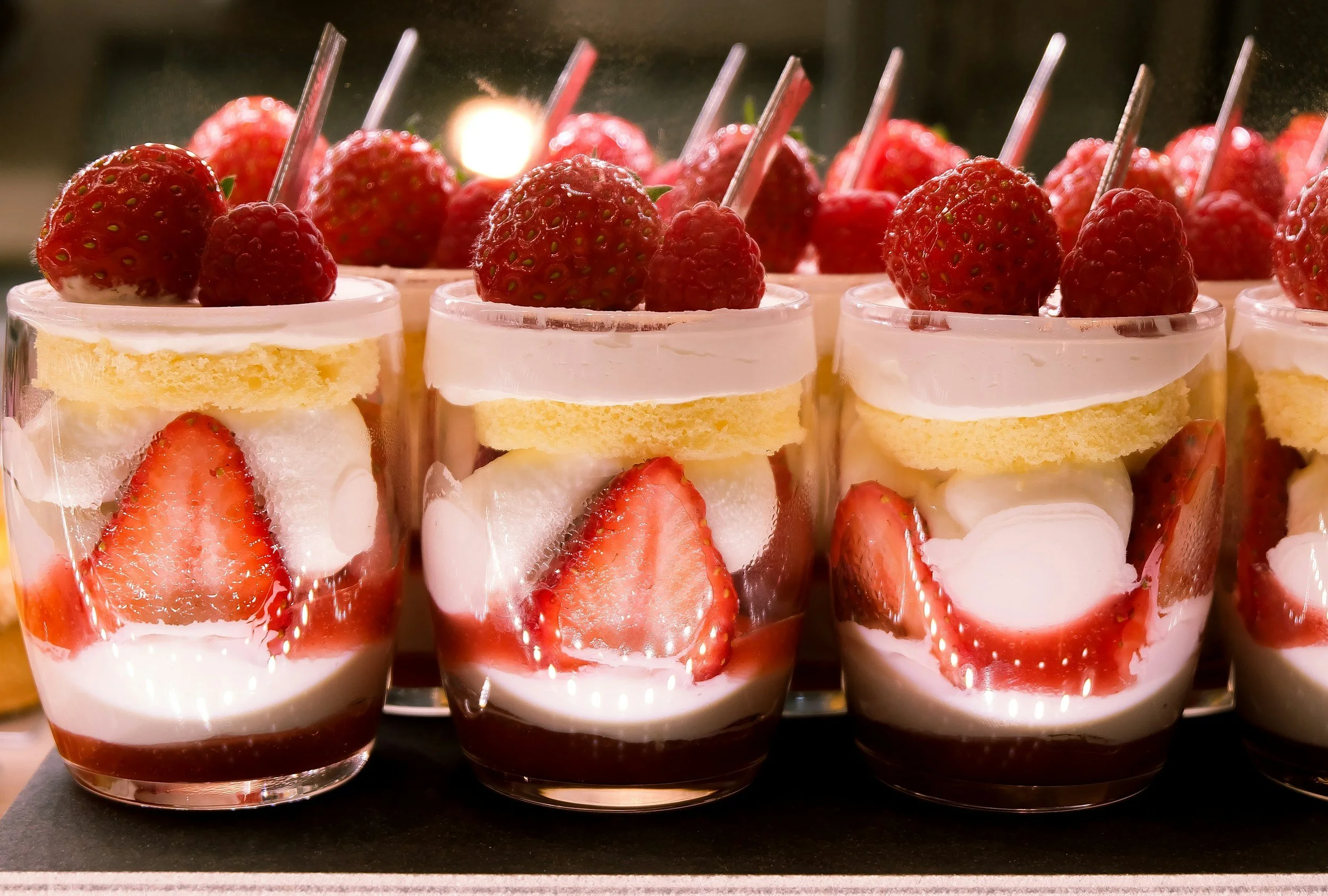 Cream parfaits in glasses topped with strawberries and slices of cake inside a display case.