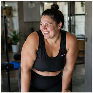 Woman with dark hair in a bun smiling, wearing a black athletic top, in a gym or fitness studio.