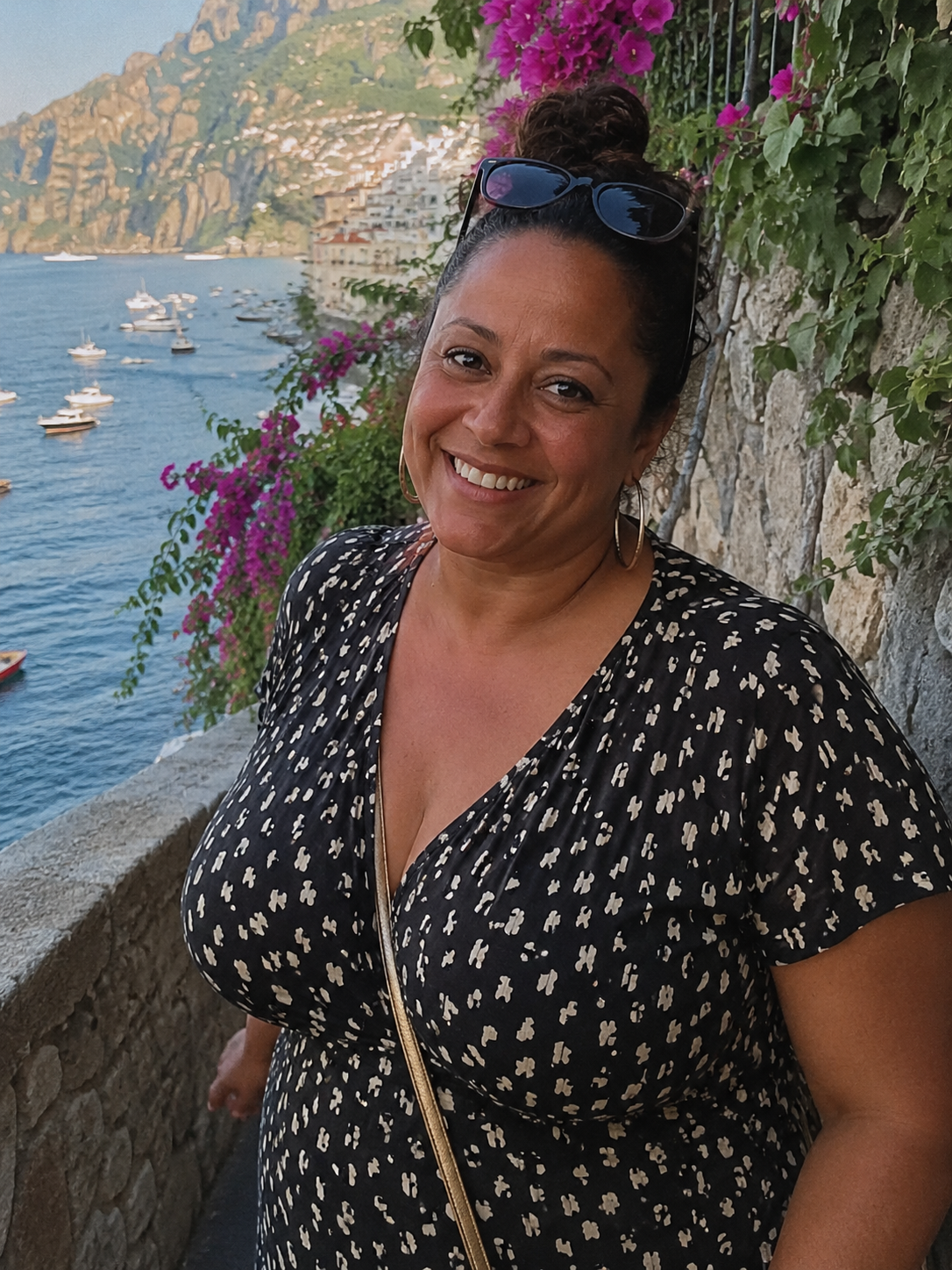 A woman smiling at the camera near a stone wall with pink flowers, a body of water, and boats in the background.