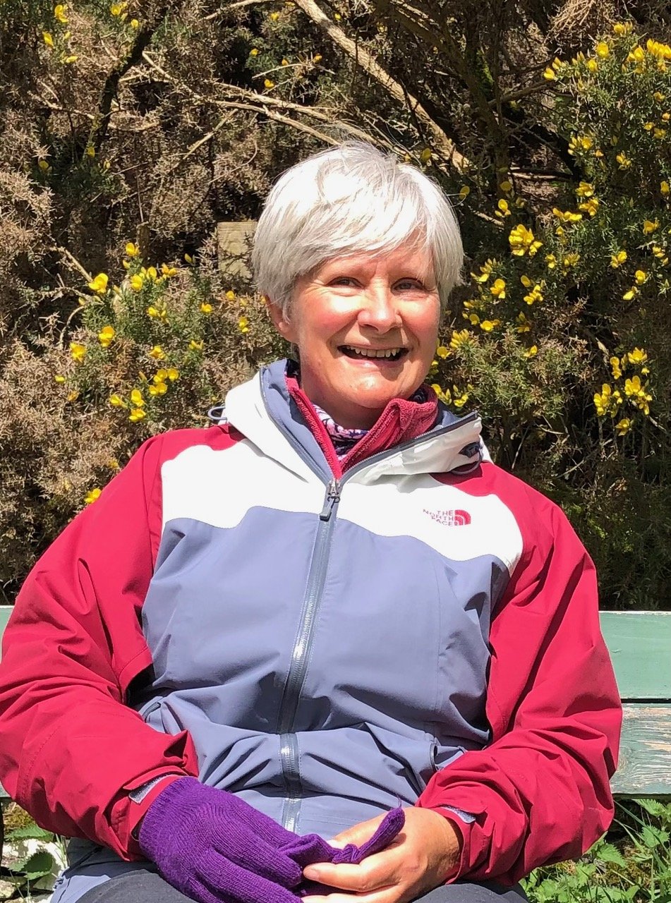 A smiling woman with short gray hair sitting on a green park bench outdoors. She is wearing a red and gray North Face jacket and purple gloves, with yellow flowering bushes and trees in the background.