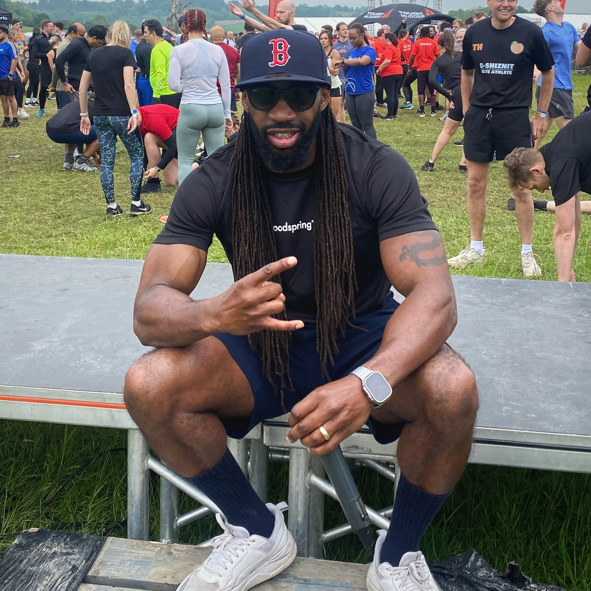 A man with long dreadlocks, sunglasses, a Boston baseball cap, a black T-shirt, and white sneakers is sitting on a barrier at an outdoor event. In the background, many people are standing, walking, and interacting on a grassy field, with some wearing athletic clothes and others in casual attire. Tents and event banners are visible.