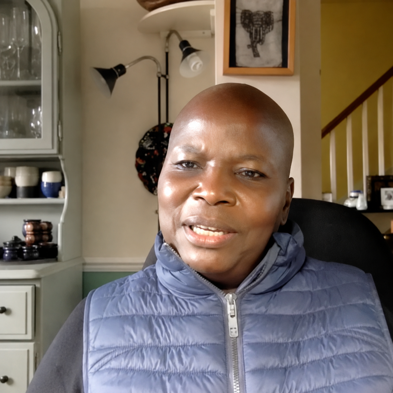 A bald man wearing a gray and blue puffer jacket sitting in a room with a cabinet, wall decorations, and a staircase in the background.