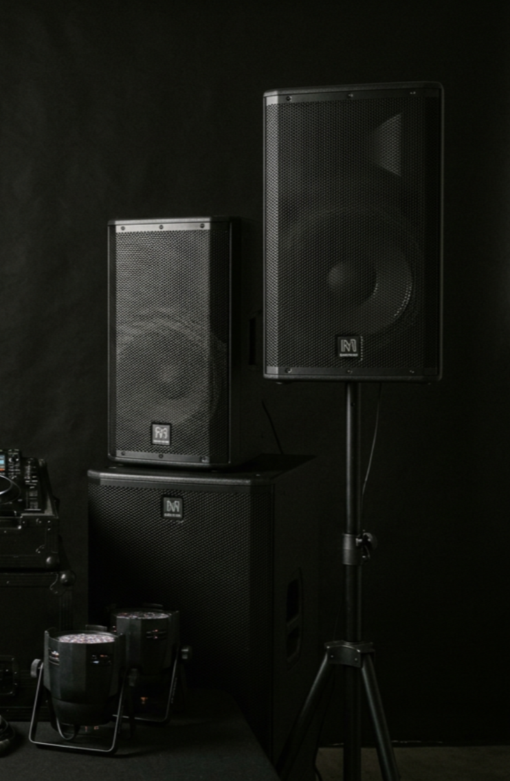 Martin speaker units, three stacked and two on stands, with festival lighting equipment on the floor, all against a black background.