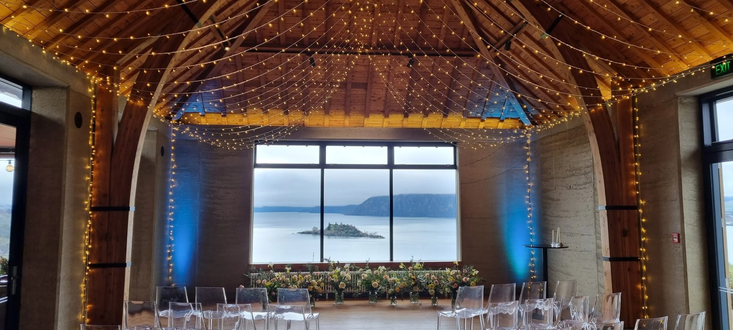 Indoor event space with high wooden ceiling and fairy lights hanging, large window with a view of the water and mountains, floral arrangements, and clear plastic chairs.