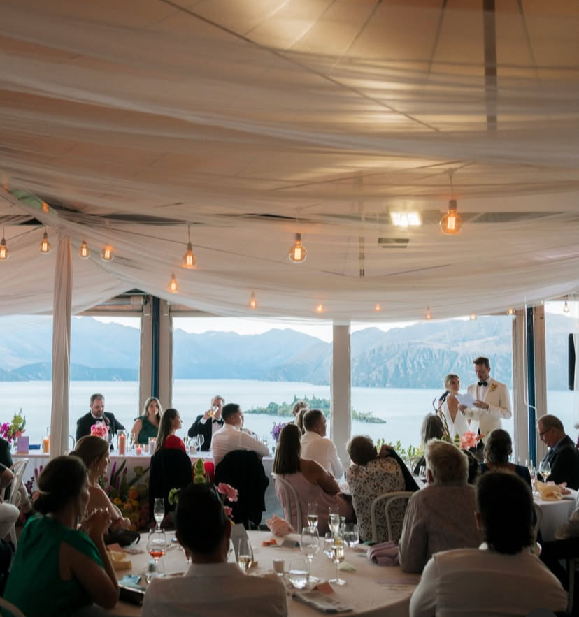 Wedding reception inside with guests seated at tables, overlooking mountains and a lake, with a bride and groom giving a toast.