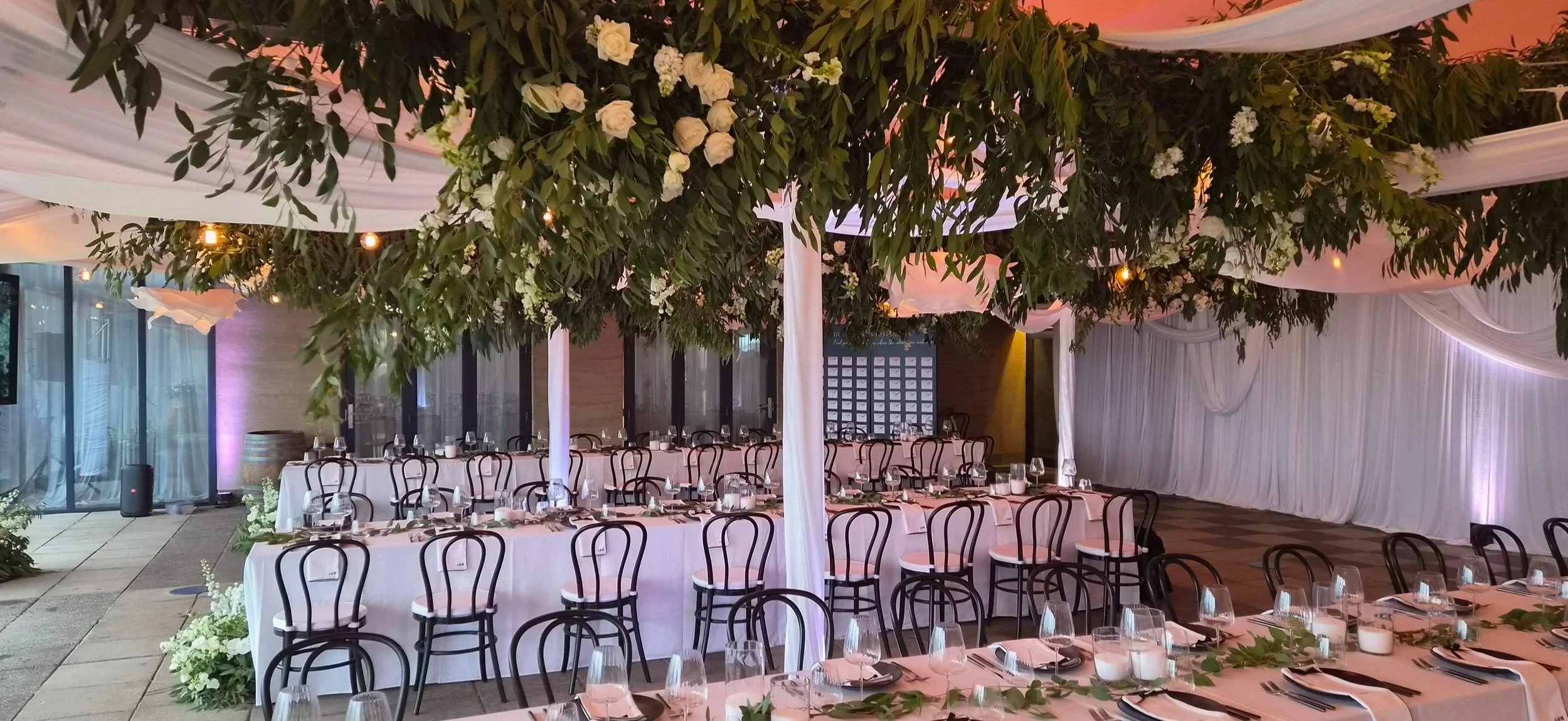 Decorated event space with hanging greenery, white flowers, draped white fabric, long banquet tables set with plates, glasses, and napkins, surrounded by black chairs, with soft lighting and purple wall lighting in the background.