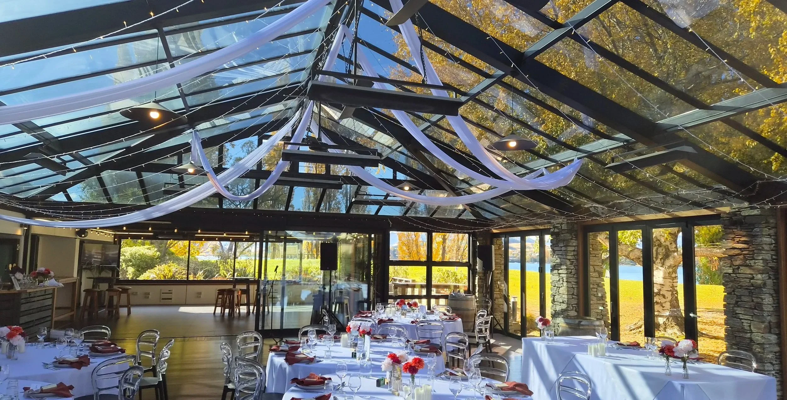 Indoor event space with tables set for a celebration, decorated with white tablecloths, napkins, and floral centerpieces. The ceiling has decorative draping and string lights, with large windows revealing a view of trees and water outside.