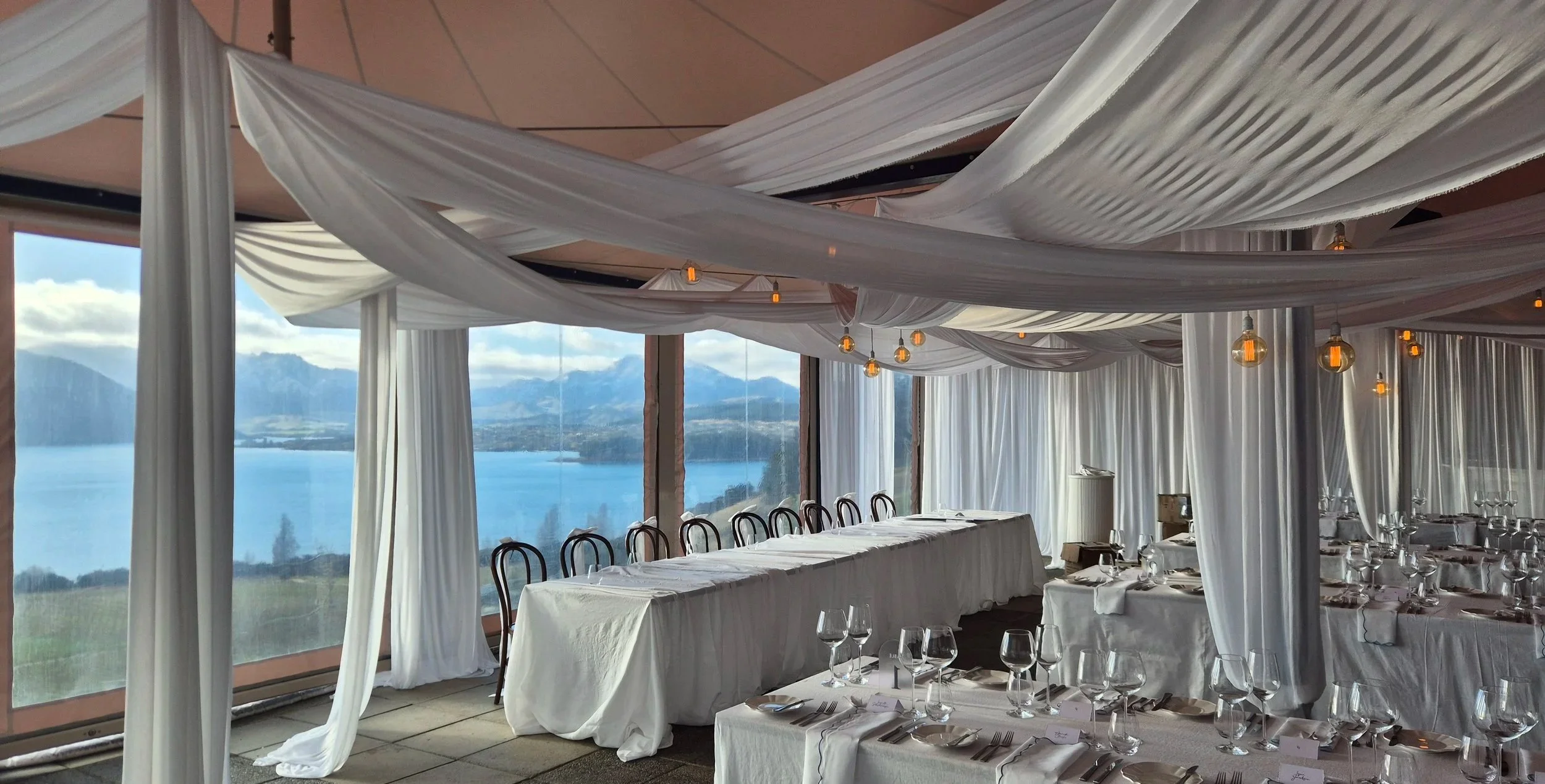Elegant event space with long dining tables covered in white tablecloths, set with glassware and silverware, and decorated with flowing white curtains and draped fabric ceiling. Large windows reveal a scenic mountain lake view.