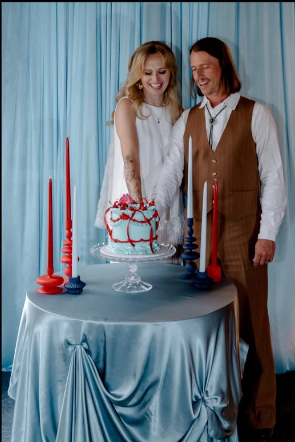 A couple is celebrating a wedding or special occasion, cutting a white cake with red and pink decorations, on a table with red and blue candles, against a blue curtain background.