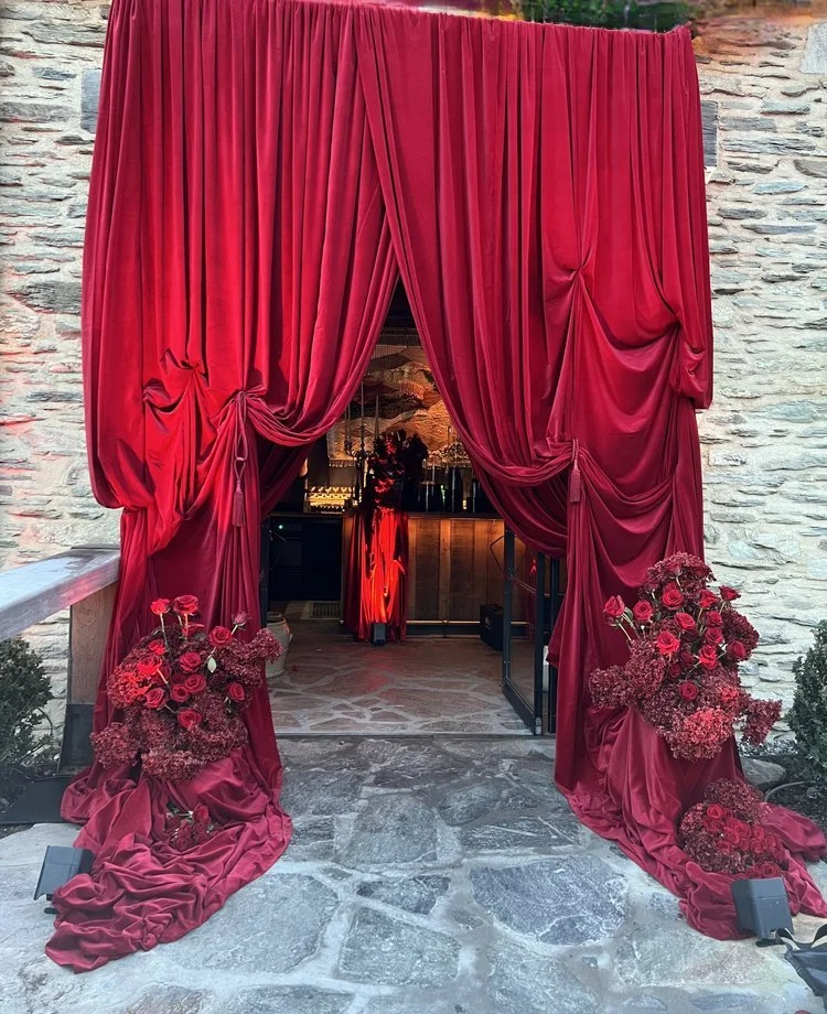 Red velvet curtains framing an entrance with floral arrangements of red roses and pink flowers on each side, leading into a decorated indoor space.