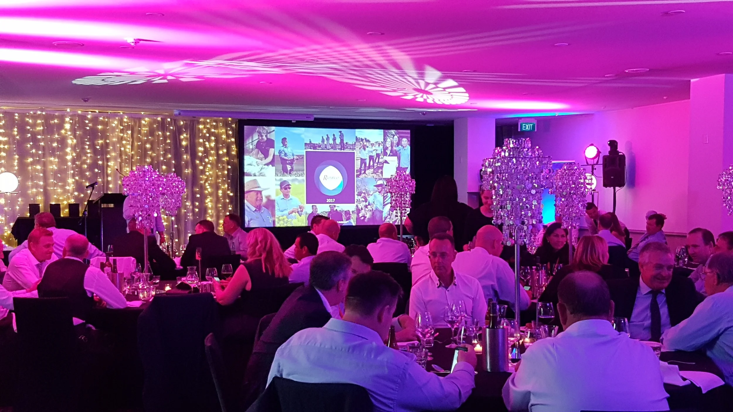 A banquet hall decorated with string lights, pink and purple lighting, and chandeliers. People are seated at tables, attending an event with a large screen displaying a collage of pictures and the year 2017.