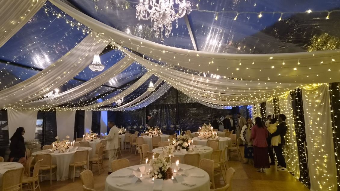 Elegant decorated event tent with draped white fabric and string lights, round tables with flower centerpieces and candles, multiple chandeliers, and guests mingling.