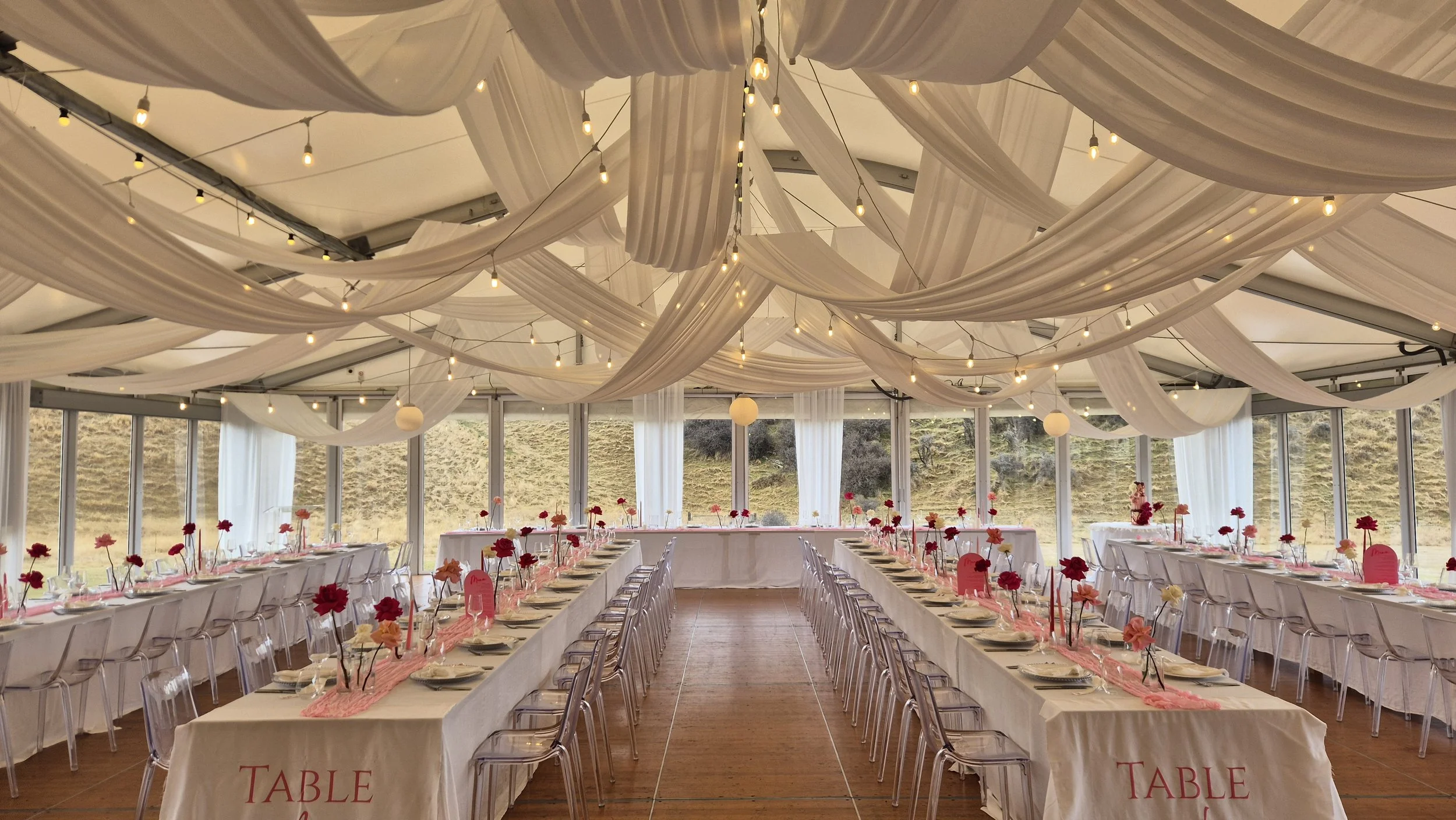 Indoor banquet hall decorated for an event with draped white fabric on the ceiling, string lights, long tables with white tablecloths, pink and red flower centerpieces, and chairs arranged along the tables.