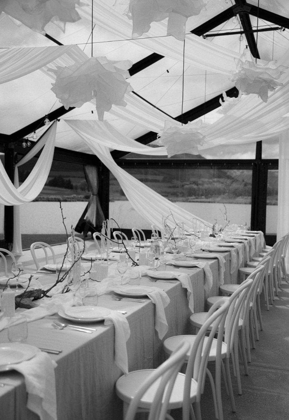 A long dining table set for a formal event in an enclosed space with large windows, decorated with hanging draped fabric and paper cloud-like decorations on the ceiling, with empty chairs arranged along the sides.
