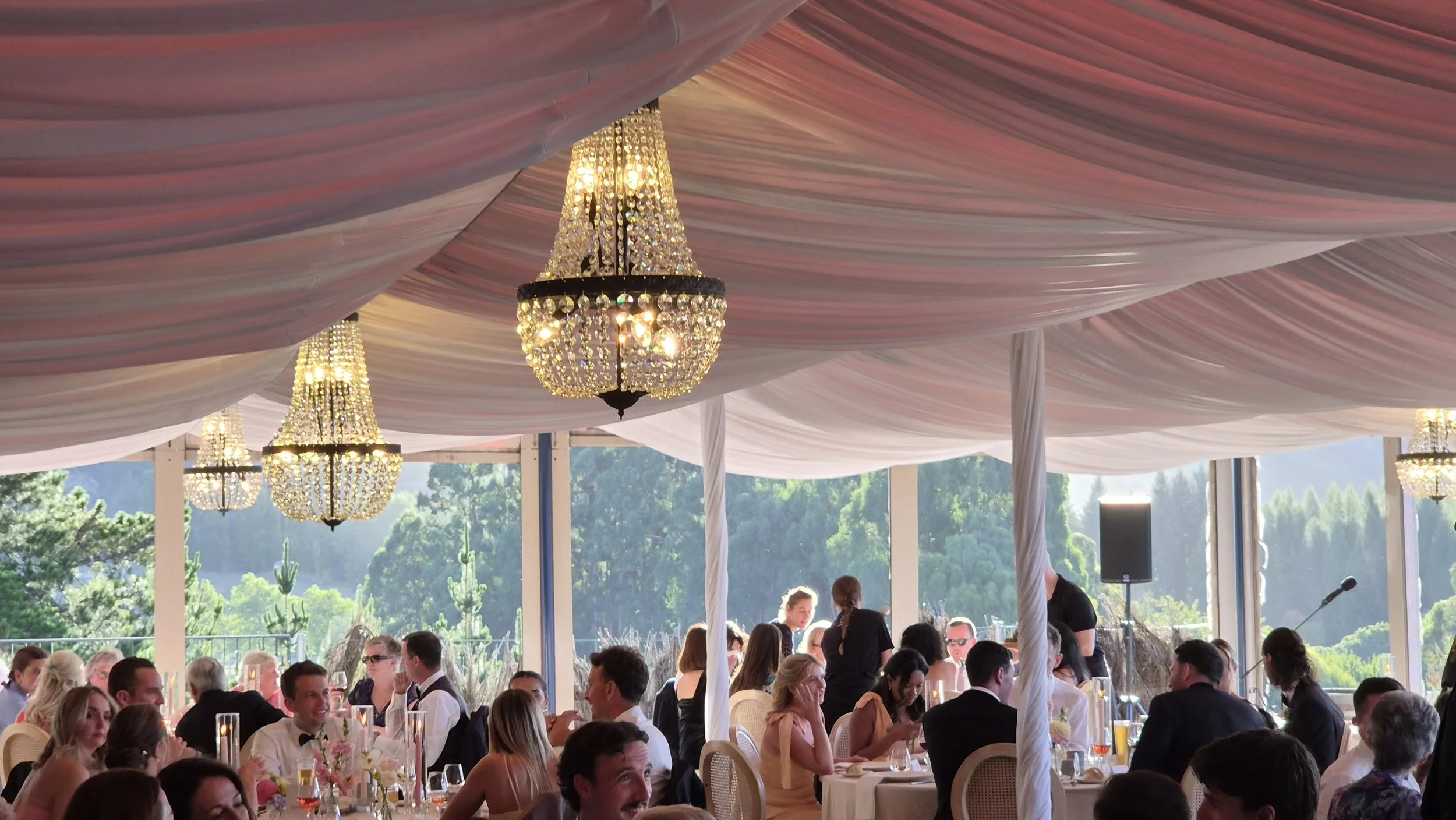 A large event space with pink and white draped fabric ceiling and multiple elegant crystal chandeliers. There are many guests seated at round tables, some standing, with a scenic outdoor view of green trees and hills in the background.