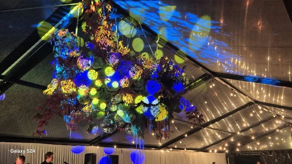 A colorful floral chandelier hanging from a ceiling with string lights in a decorated event tent.