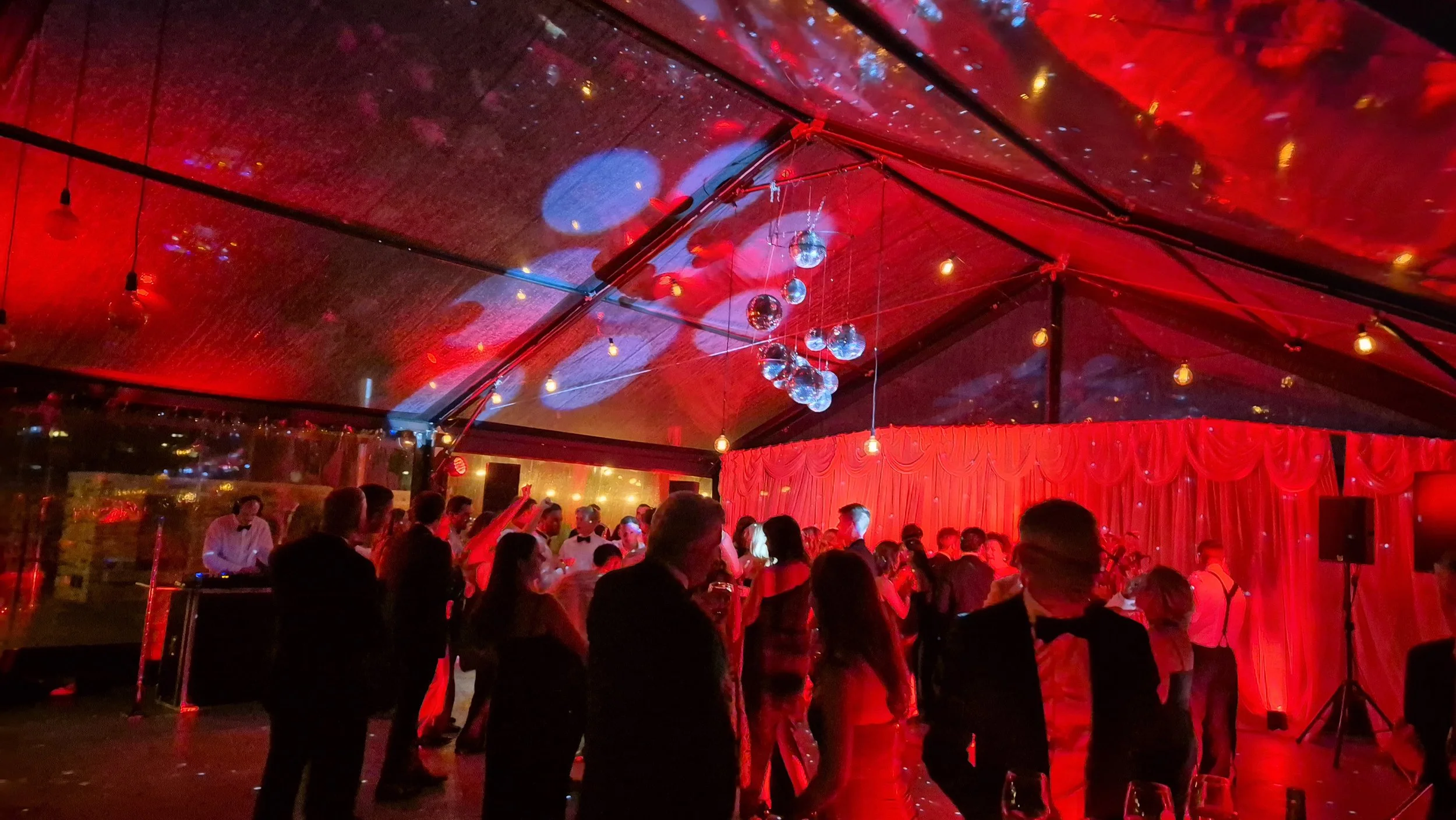 People dancing and socializing at a lively party or event inside a red-lit venue with disco balls and string lights on the ceiling.