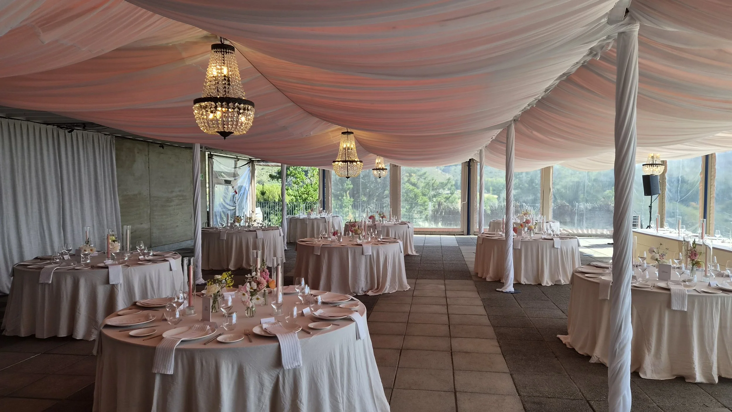 Elegant wedding reception setup inside a tent with draped pink fabric on the ceiling, chandeliers, round tables with pink tablecloths, floral centerpieces, white plates, glasses, and cutlery arranged for guests.