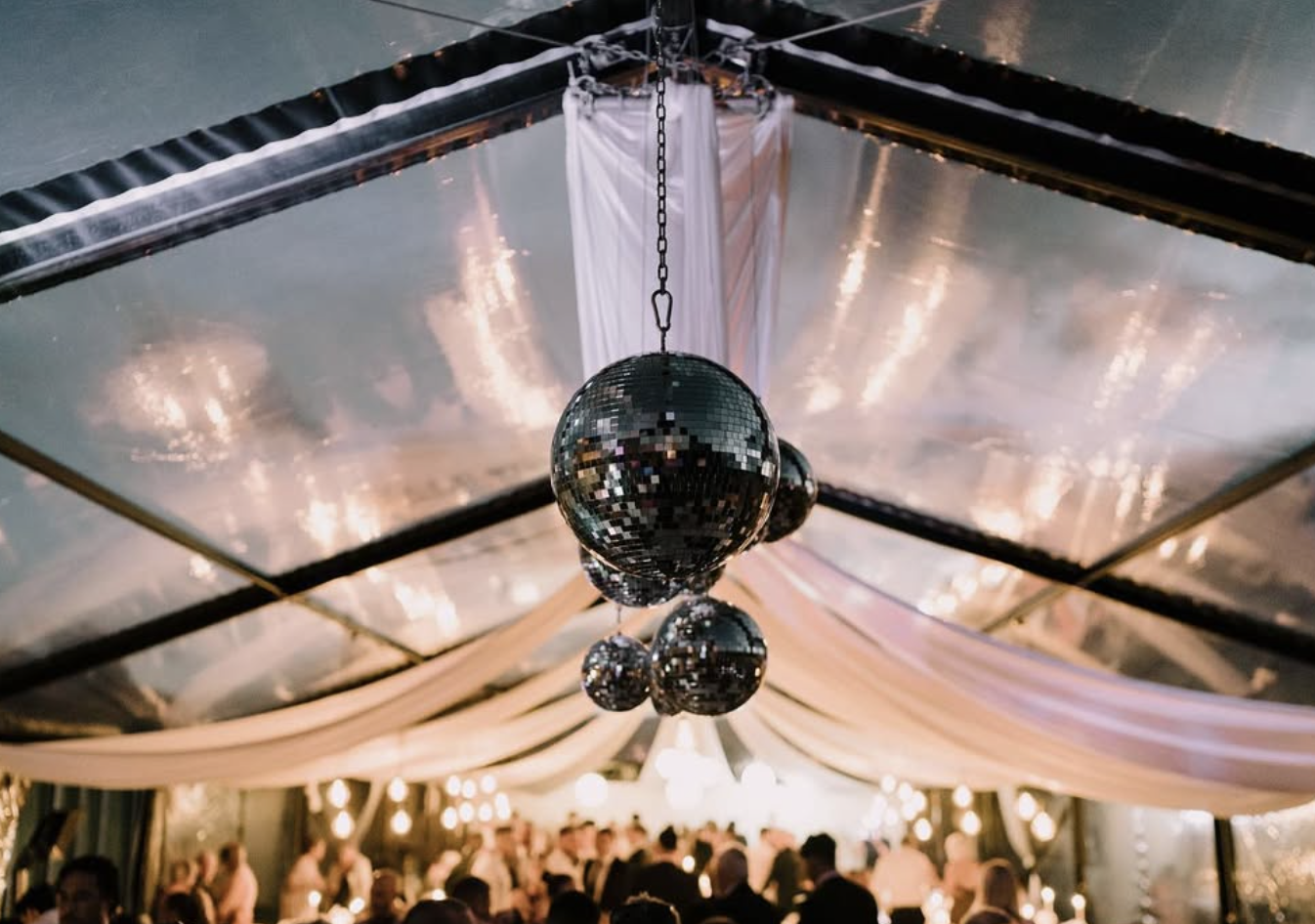 Hanging disco ball decorations in a decorated event venue with soft lighting in a clear marquee, with people socialising below.