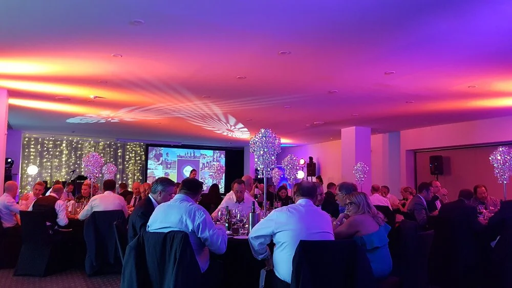 People attending a formal event or banquet in a decorated banquet hall with purple and pink lighting, floral centerpieces, and a large screen at the front.
