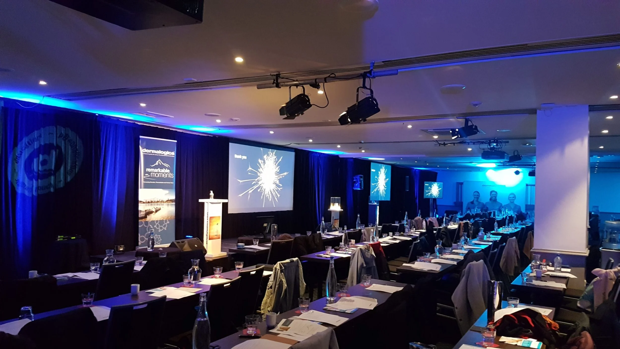 Conference room set up for a presentation with blue lighting, multiple projection screens, and tables arranged with water bottles and notes.